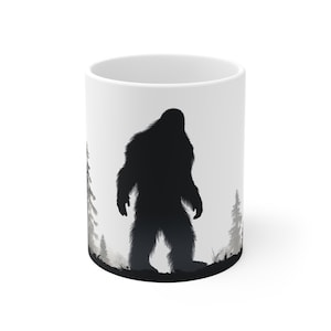 May include: White ceramic mug featuring a black silhouette of Bigfoot standing in a forest scene. The design includes trees and a misty background, creating a mysterious and outdoorsy theme. Perfect for coffee or tea.