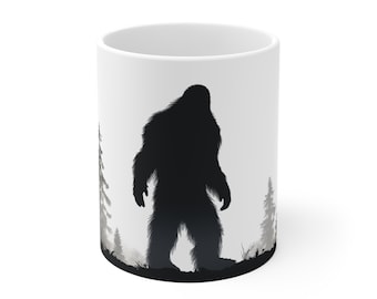 May include: White ceramic mug featuring a black silhouette of Bigfoot standing in a forest scene. The design includes trees and a misty background, creating a mysterious and outdoorsy theme. Perfect for coffee or tea.