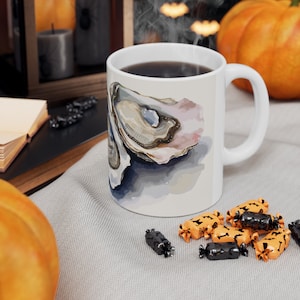 May include: White ceramic mug featuring a detailed illustration of an oyster shell with a pearl-like interior. The mug is filled with dark liquid, possibly coffee or tea. The design includes shades of beige, pink, and blue. The mug has a white handle.