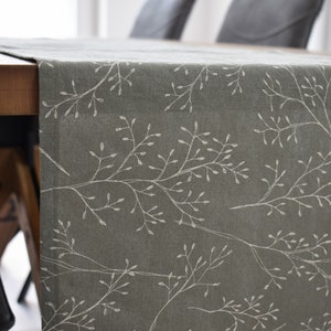 Könnte beinhalten: Ein grüner Tischläufer aus Leinen mit einem weißen Blumenmuster. Der Läufer liegt auf einem Holztisch mit zwei Stühlen.