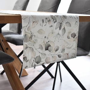 Könnte beinhalten: Ein weißer Tischläufer mit einem Aquarell-Blumendesign mit grauen und rosafarbenen Blättern und Blüten. Der Läufer liegt auf einem Holztisch mit zwei grauen Stühlen.