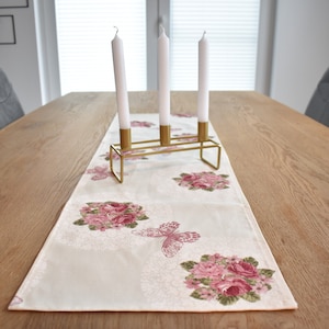 Könnte beinhalten: Ein weißer Tischläufer mit einem Blumendesign mit rosa Rosen und Schmetterlingen. Ein goldfarbener Kerzenhalter aus Metall mit drei weißen Kerzen steht auf dem Läufer.