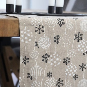 Könnte beinhalten: Beiger Tischläufer mit einem sich wiederholenden Muster aus weißen und schwarzen Schneeflocken, Sternformen und stilisierten floralen Designs. Der Läufer liegt auf einem Holztisch, mit schwarzen und weißen Kerzen im Hintergrund.