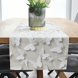 Könnte beinhalten: Ein beiger Tischläufer mit einem weißen Schmetterlingsmuster. Der Läufer liegt auf einem Holztisch mit einer Vase mit weißen Blumen in der Mitte.
