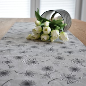 Könnte beinhalten: Ein grauer Tischläufer mit einem Löwenzahnmuster. Der Läufer liegt auf einem Holztisch mit einem Strauß weißer Tulpen in einer Glasvase.