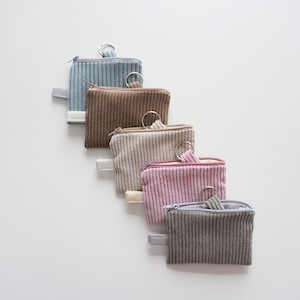 Könnte beinhalten: Fünf kleine Cord-Münzbörsen in einer Reihe, jede in einer anderen Farbe: Hellblau, Braun, Beige, Rosa und Grau. Jede Geldbörse hat einen Reißverschluss, einen Metallring und eine kleine Stoffschlaufe.