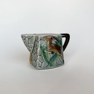 May include: A ceramic creamer pitcher featuring a raised relief design of a brown gazelle among green leaves against a gray stone-textured background. The pitcher has a dark brown handle and a distinct pouring spout, showcasing a glossy finish.