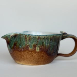 May include: A ceramic creamer pitcher featuring a rounded body with a textured brown base and a glossy green and blue drip glaze pattern on the upper portion. The vessel includes a small pouring spout on one side and a curved handle on the opposite side.