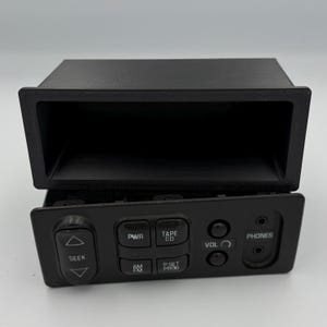 Puede incluir: Autorradio negro con una abertura rectangular sobre un panel de control. El panel de control tiene botones etiquetados SEEK, PWR, TAPE CD, AM FM, P SET PROG, VOL y PHONES. La radio está diseñada para encajar en el salpicadero de un coche.