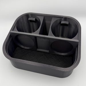 Puede incluir: Un organizador de consola de coche negro, impreso en 3D, con dos portavasos y un compartimento de almacenamiento. El organizador tiene una base texturizada y una forma rectangular con esquinas redondeadas. Los portavasos tienen pequeñas pestañas.