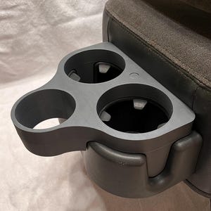 以下が含まれることがあります： グレーのプラスチック製のカップホルダーで、カップを置くスロットが3つあります。カップホルダーはグレーの布張りの座席に取り付けられています。