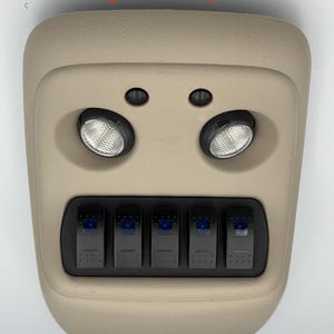 Puede incluir: Consola de techo de color beige con dos luces circulares y cinco aberturas rectangulares para interruptores. El texto "Designed to Stay Put" está arriba y "Switches not included" abajo.