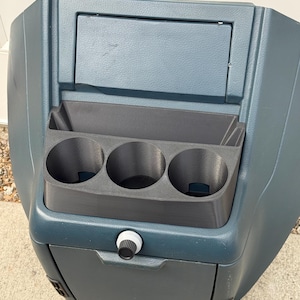 May include: A dark blue automotive center console featuring a textured plastic finish, a rectangular storage compartment with a lid, and a black insert containing three circular cup holders. A small black and white knob is centered on the front.
