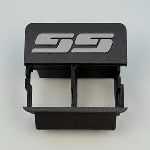 Puede incluir: Inserto de consola de coche de plástico negro con las letras "SS" en gris. El inserto tiene dos compartimentos y una parte superior rectangular con las letras. El artículo está diseñado para encajar en la consola de un coche.