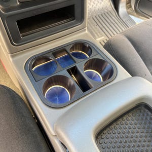 Puede incluir: Un portavasos de plástico negro para la consola central de un vehículo, con cuatro aberturas circulares y revestimientos de goma azul texturizada. El inserto está colocado en una consola gris, debajo de una unidad de control de audio.