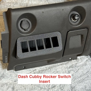 May include: A dark gray dash cubby rocker switch insert with five rectangular openings. The insert is part of a larger dashboard component, featuring a circular opening, a cigarette lighter, and a small storage compartment. The text "Dash Cubby Rocker Switch Insert" is visible.