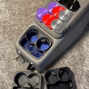 Puede incluir: Una consola de coche gris con varios insertos para portavasos y soportes magnéticos para teléfono de colores. Los insertos para portavasos son negros y grises. Los soportes magnéticos son morados, rojos, plateados y negros.