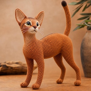 May include: A crocheted cat figurine in shades of orange and brown. The cat has large, pointed ears, a long neck, and a slender body. Its tail is upright, with a darker brown tip. The cat is standing on all fours, with its eyes looking upwards.
