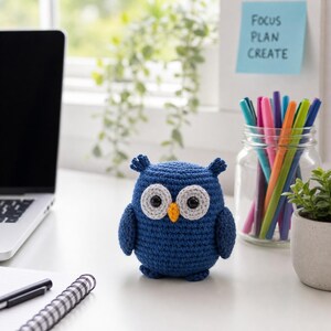 Puede incluir: Un b&uacute;ho azul tejido a mano con ojos grises y blancos y un pico naranja est&aacute; sobre un escritorio blanco. Tambi&eacute;n hay un port&aacute;til, bol&iacute;grafos y una peque&ntilde;a planta en el escritorio. Una nota adhesiva dice "Focus Plan Create".