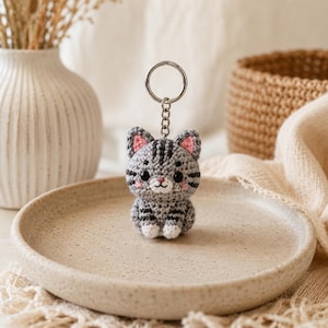 May include: A gray and white crocheted cat keychain featuring dark gray stripes, black bead eyes, pink stitched inner ears, and a small pink nose. The cat figure is attached to a silver-colored metal split ring by a short chain.