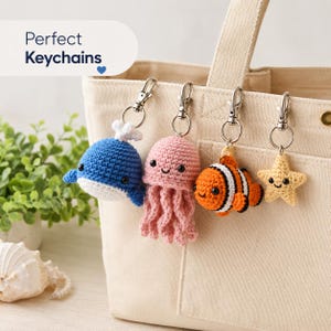 May include: Four crochet keychains attached to a beige canvas bag. The charms include a blue whale with a white spout, a pink jellyfish with dangling tentacles, an orange and white clownfish, and a yellow star, all featuring black bead eyes.