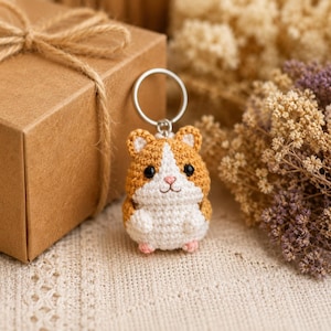 May include: A small, crocheted hamster keychain featuring a round body with tan and white yarn, black bead eyes, and a pink stitched nose. The hamster is attached to a silver-toned metal split ring, creating a compact accessory for keys.