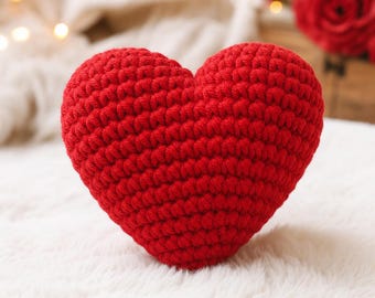 May include: A handmade, red crocheted heart. The heart is centrally positioned on a white, textured surface. The yarn is a vibrant red, and the stitches are clearly visible, giving the heart a soft, tactile appearance. The background is blurred, with hints of white and red.