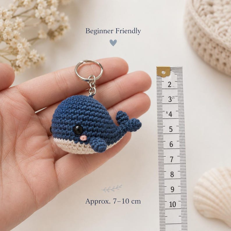 May include: A dark blue and cream crochet whale keychain with a metal ring. The whale features a small black eye and a pink cheek detail. A ruler is positioned next to the whale, indicating a size range of approximately 3 to 4 inches.