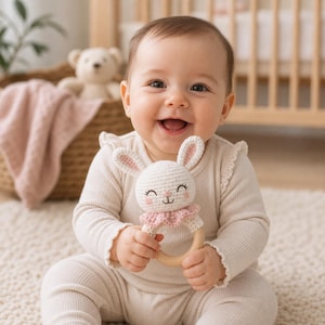 May include: A smiling baby holds a white and pink crochet bunny rattle with a wooden ring. The baby wears a light beige onesie. A basket with a pink blanket and a teddy bear are in the background.