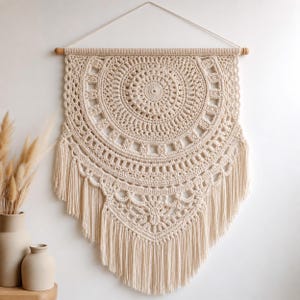 May include: Cream-colored, handmade macrame wall hanging. The circular design features intricate crochet patterns and a fringe at the bottom. It is suspended from a wooden dowel with a natural fiber cord. This bohemian-style wall decor adds texture and warmth to any space.
