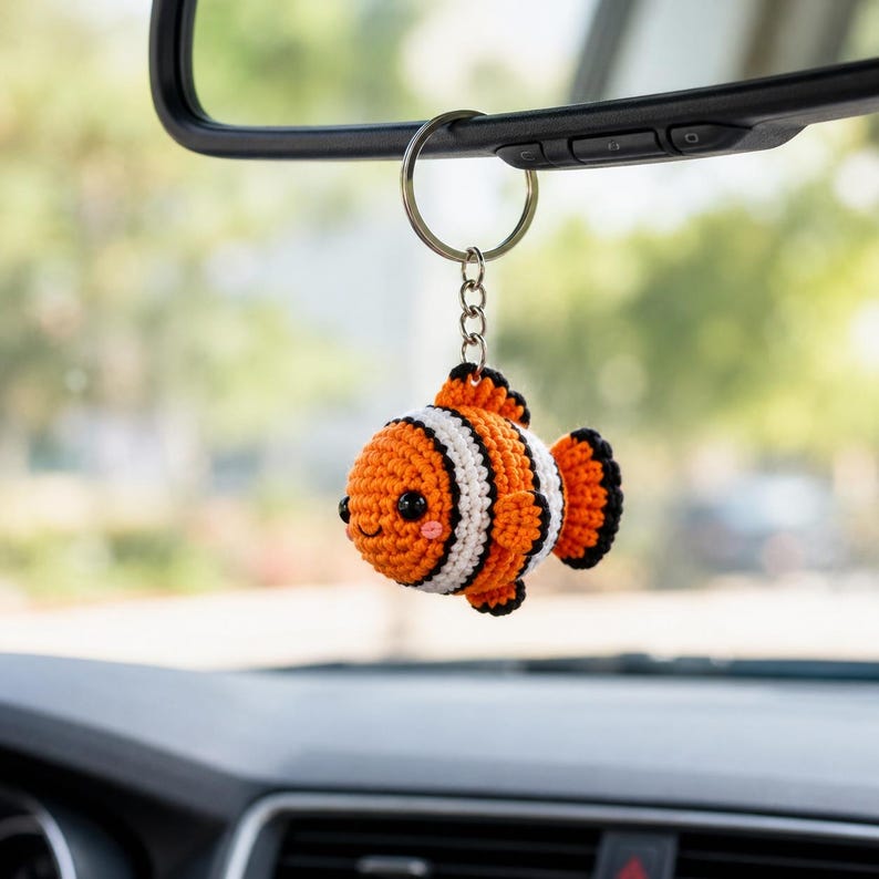 May include: A crocheted clownfish keychain hanging from a car rearview mirror. The fish is orange with white and black vertical stripes, featuring black bead eyes, a small stitched smile, and pink blush accents on its round, textured body.