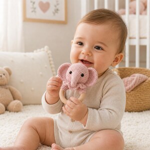 May include: A smiling baby holds a pink crochet elephant rattle with a wooden ring. The rattle features a detailed elephant head with small black eyes and large ears. The baby is wearing a light-colored onesie and sitting on a white rug.