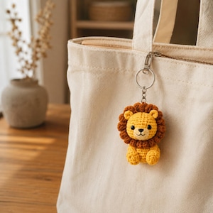 May include: A crochet lion keychain featuring a yellow body and a dark brown mane, attached to a silver-toned metal keyring. The lion has small rounded ears, black bead eyes, and a stitched nose and mouth, hanging from a cream-colored canvas tote bag.