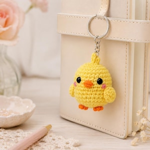 May include: A small, round, yellow crocheted chick keychain featuring black plastic eyes, a small orange beak, tiny orange feet, and pink embroidered cheeks. The chick is attached to a silver-toned metal split ring and a short chain.