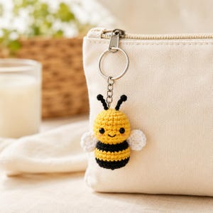 May include: A small, crocheted bee keychain featuring a yellow and black striped body, two white wings, and black antennae. The bee is attached to a silver-toned metal split ring and chain, which is fastened to the zipper pull of a beige canvas pouch.