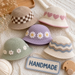 Op de afbeelding: Vijf handgemaakte gehaakte vissershoeden in verschillende kleuren en patronen. De hoeden hebben een dambordpatroon, roze harten, madeliefjes en golvende strepen. Een bord met de tekst "HANDMADE" is ook zichtbaar.