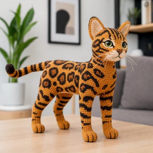 Op de afbeelding: Een gehaakte kattenfiguur in een staande pose. De kat heeft een bruin lichaam met zwarte vlekken en strepen, groene ogen en lange snorharen. De kat is ongeveer 30 cm hoog en staat op een houten tafel.