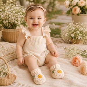 May include: A cream-colored knit romper featuring ruffled shoulder straps and two small wooden buttons at the chest. The outfit is paired with matching cream-colored crochet booties, each decorated with a white and yellow daisy flower motif.