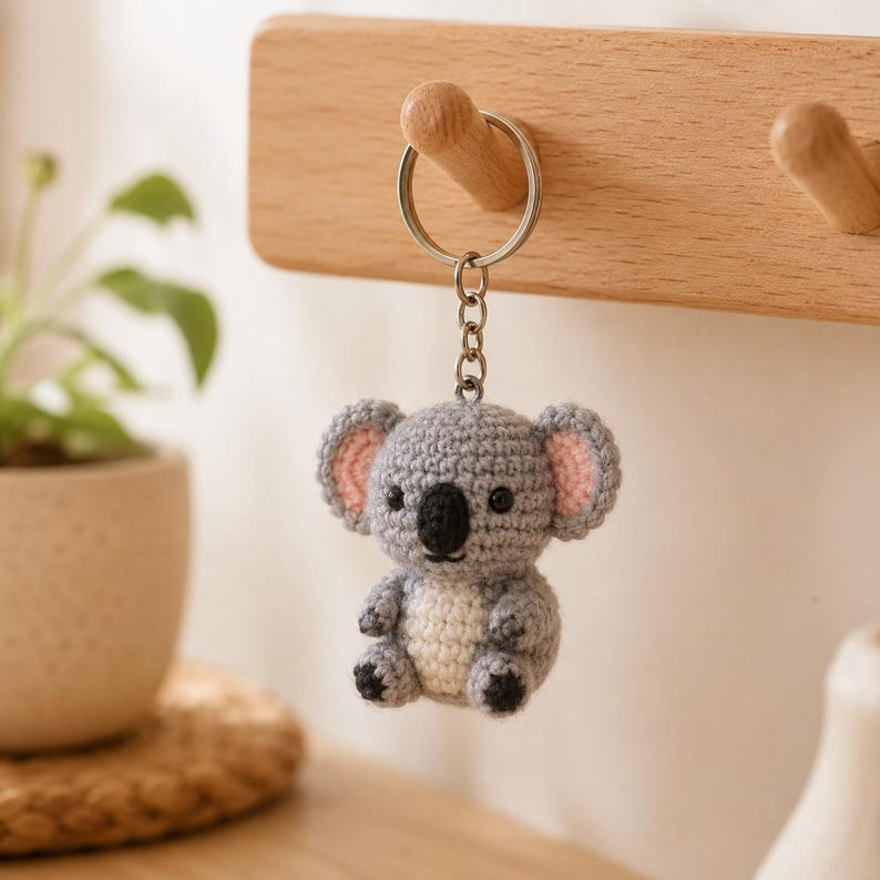 May include: A small, crocheted koala keychain featuring a grey body, a cream-colored belly, and pink inner ears. The koala has black embroidered eyes and a black nose, attached to a silver-toned metal split ring by a short metal chain.