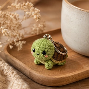 May include: A small, crocheted turtle keychain featuring a light green head and body, a dark brown textured shell, black bead eyes, and small pink stitched cheeks. The turtle is attached to a silver-toned metal split ring keychain.
