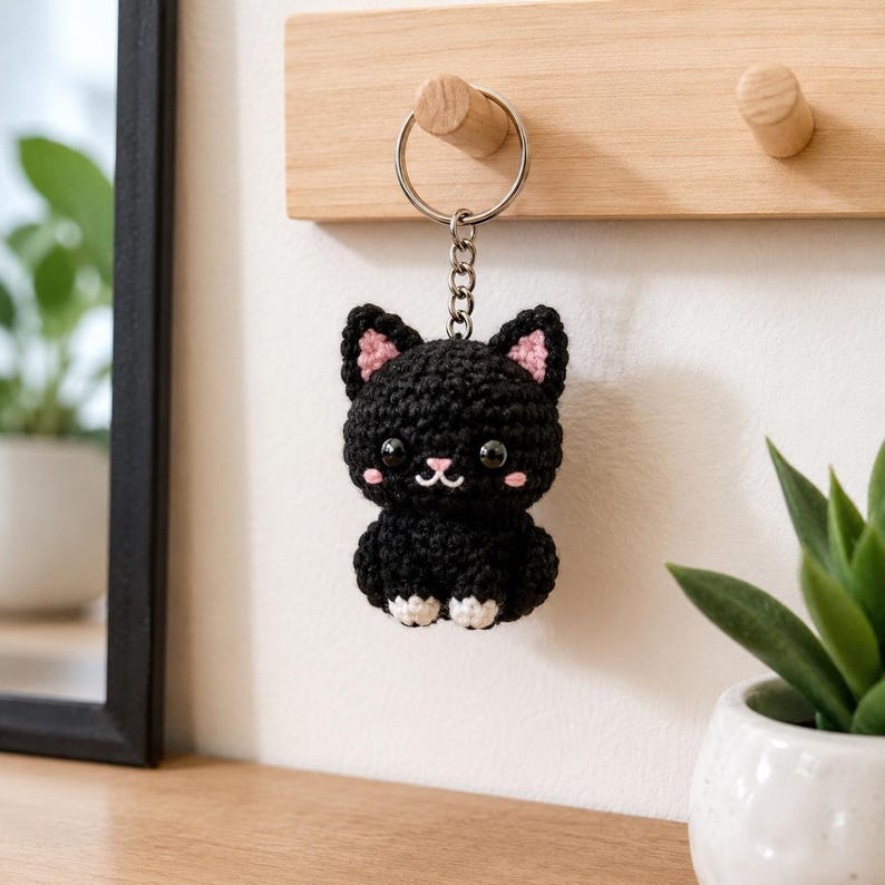May include: A black crochet cat keychain featuring a round head, pointed ears with pink inner details, large black plastic eyes, a small pink stitched nose, and white paws. The plush cat is attached to a silver metal split ring and chain.