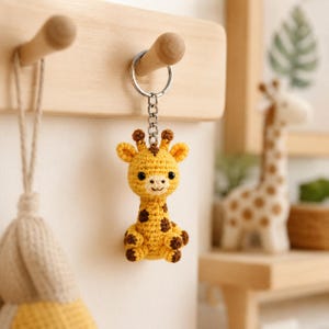 May include: A small, handmade crochet giraffe keychain featuring a yellow body with brown spots, a light-colored muzzle, and two small horns. The plush giraffe is attached to a silver-toned metal split ring by a short chain, hanging vertically.