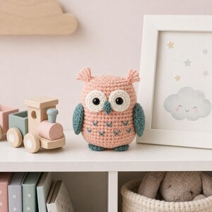 Puede incluir: Un juguete de b&uacute;ho tejido a mano en rosa suave con detalles blancos y turquesa. El b&uacute;ho tiene grandes ojos negros y est&aacute; colocado en una estanter&iacute;a blanca con un tren de madera y una imagen enmarcada de una nube y estrellas. Decoraci&oacute;n de guarder&iacute;a.