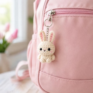May include: A small, cream-colored crocheted rabbit keychain with long ears featuring pink inner details, black bead eyes, and a tiny embroidered nose. The rabbit is attached to a silver-toned metal chain and ring, hanging from a pink fabric backpack.