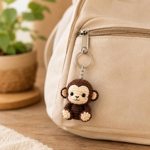 May include: A small, crocheted monkey keychain attached to a beige canvas backpack. The monkey features dark brown yarn for the body and head, with a lighter cream-colored face, ears, and paws, plus two small pink blush marks on its cheeks.