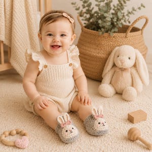 May include: A pair of grey crochet baby booties featuring a white bunny face design with pink ears and a small pink bow. The booties are crafted with a soft texture and a rounded toe shape, designed to fit comfortably on small feet for daily wear.