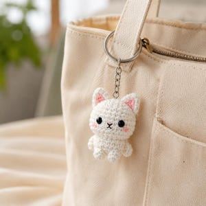 May include: A white crocheted cat keychain featuring a small, stuffed figure with pink inner ears, black bead eyes, and a stitched nose. The cat is attached to a silver-toned metal split ring and chain, which is hooked onto a beige canvas tote bag.