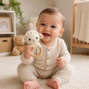 May include: A smiling baby holds a crochet lamb rattle with a wooden ring. The rattle is cream-colored with black eyes and a pink mouth. The baby wears a cream-colored waffle-knit onesie and sits on a cream-colored rug.