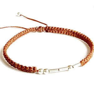 May include: A handmade macrame bracelet in shades of brown, featuring a woven design. The bracelet includes silver-colored beads and a sliding knot closure for adjustable sizing. The overall design is simple and elegant.