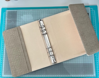May include: An open, beige-colored binder with a silver metal ring mechanism. The cover and spine feature a textured, faux-leather material in a light taupe color. The binder is lying on a teal cutting mat with grid lines.